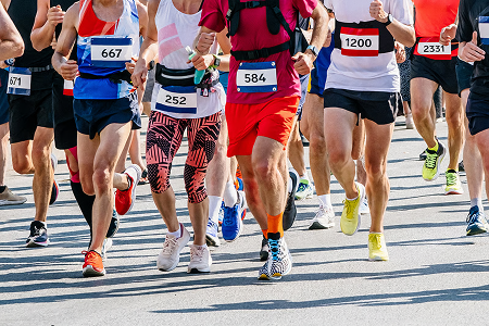 Runners in colorful athletic gear race closely together on a sunlit street, numbered bibs visible, conveying energy and competition in a marathon event.