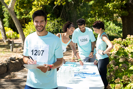 A smiling man with race bib 1207 writes on a clipboard in a sunny park. Behind him, three people gather around a table with water bottles and medals.