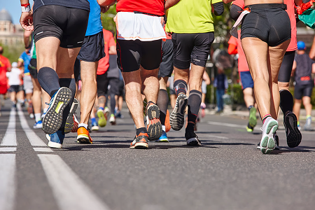 A group of runners in colorful athletic gear jog on a sunlit road during a marathon. The focus is on their legs and running shoes, conveying energy and motion.