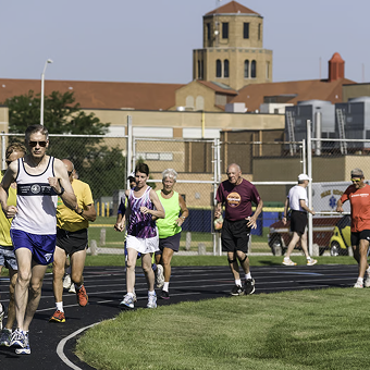 People of varying ages jogging on a sunny day at a track by a school. The mood is energetic, with buildings and a tower in the background.