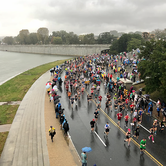 Aerial view of a large group of runners in a marathon on a rainy day. Participants in colorful gear navigate a wet road beside a river and lush trees.