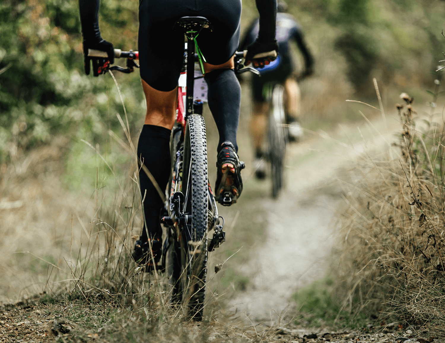 Two cyclists in black gear ride mountain bikes on a narrow dirt trail surrounded by dry grass, conveying adventure and focus.