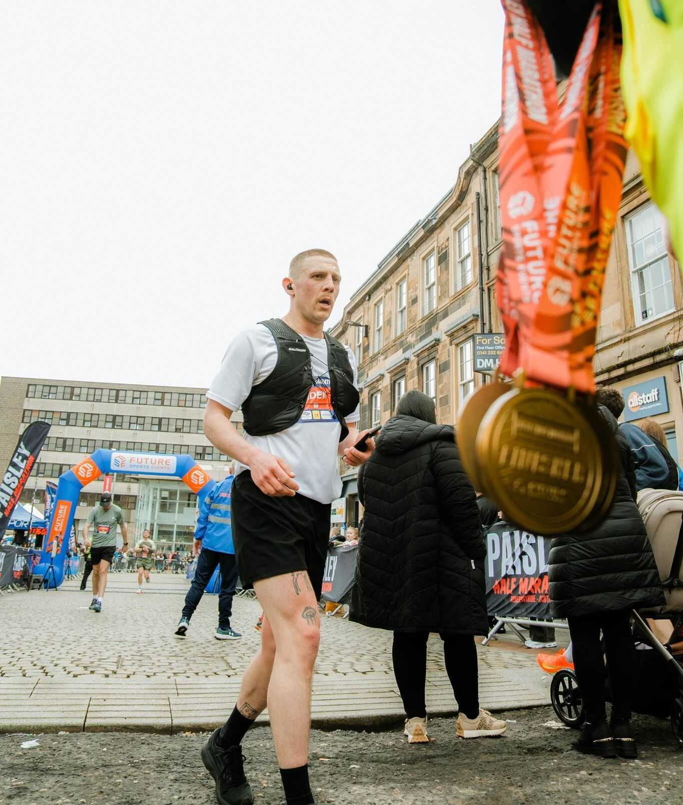 Runner nearing a marathon finish line, wearing a race bib and hydration vest. A hand holds medals in the foreground. Urban backdrop with onlookers.wearing a race bib and hydration vest. A hand holds medals in the foreground. Urban backdrop with onlookers.