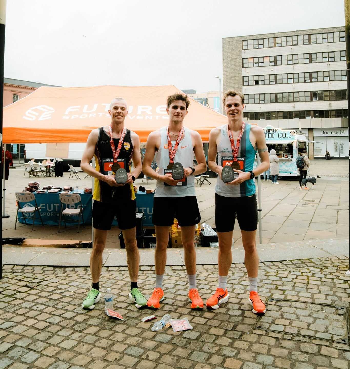 Three athletes in sports attire stand proudly holding trophies, each wearing medals around their necks. They are under an orange tent with a city backdrop.